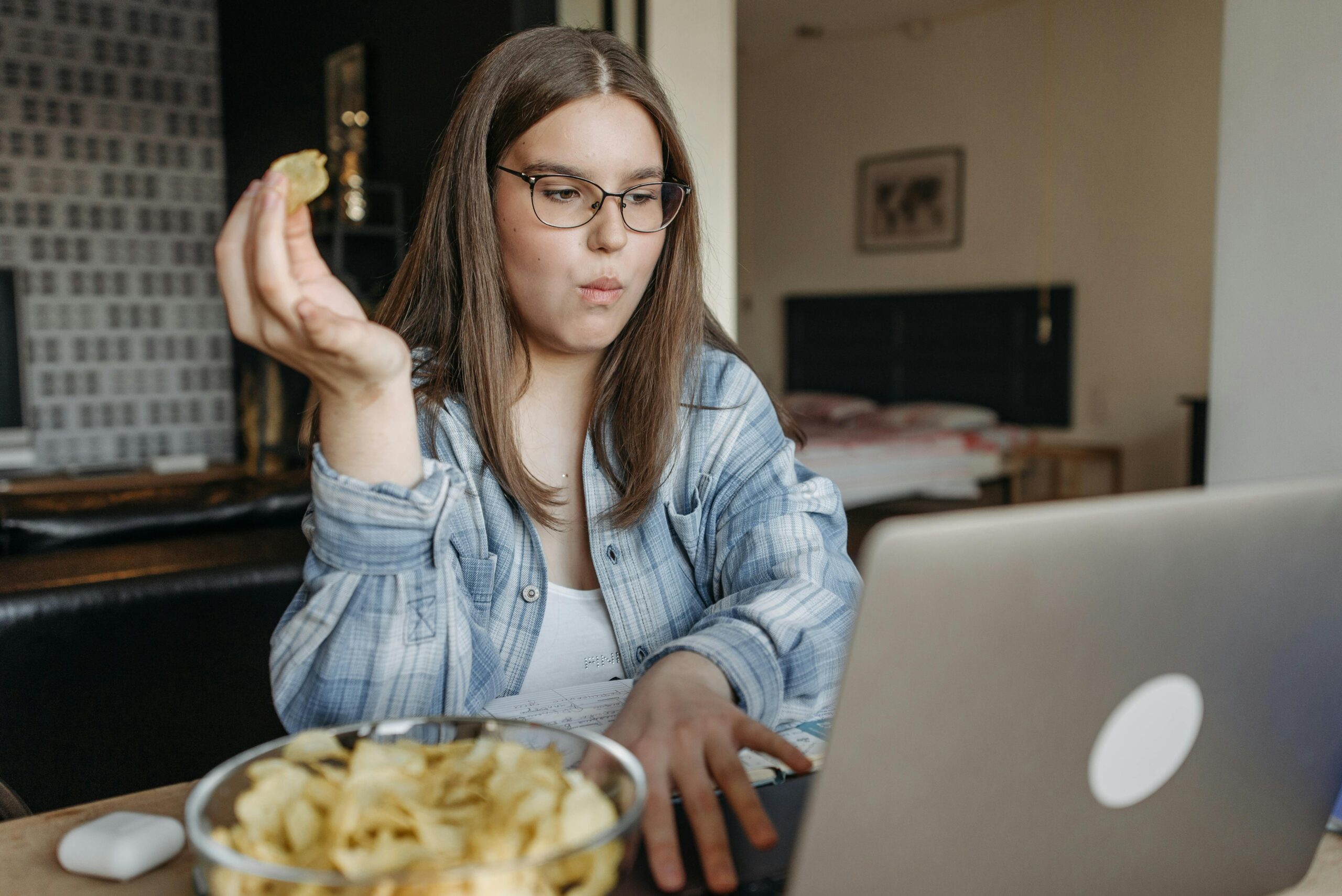 Woman in glasses working on laptop while eating chips in a cozy home setting.